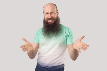 What do you need? Portrait of angry middle aged bald man with long beard in light green t-shirt standing, looking at camera with raised arms and asking. indoor studio shot isolated on grey background