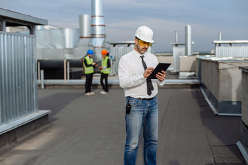 On the rooftop of construction site man engineer make a conversation with some worker using the tablet two workers are seen in the background