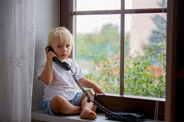 Adorable little boy, sitting on window shield, speaking on old phone on a rainy day,
