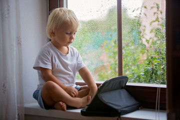 Adorable little boy, sitting on window shield, playing on tablet on a rainy day