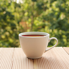 white cup with coffee on a light wooden table against a green summer garden, close-up, copy space, square