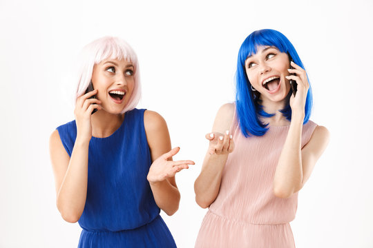 Portrait of two alluring women wearing blue and pink wigs looking at each other while talking on cellphones