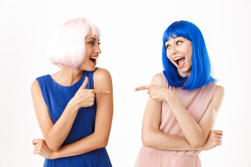 Portrait of two delighted women wearing blue and pink wigs pointing fingers at each other