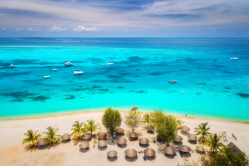 Aerial view of umbrellas, palms on the sandy beach of Indian Ocean at bright sunny day. Summer holiday in Africa. Tropical seascape with green palm trees, parasols, boats, yachts, blue water. Top view