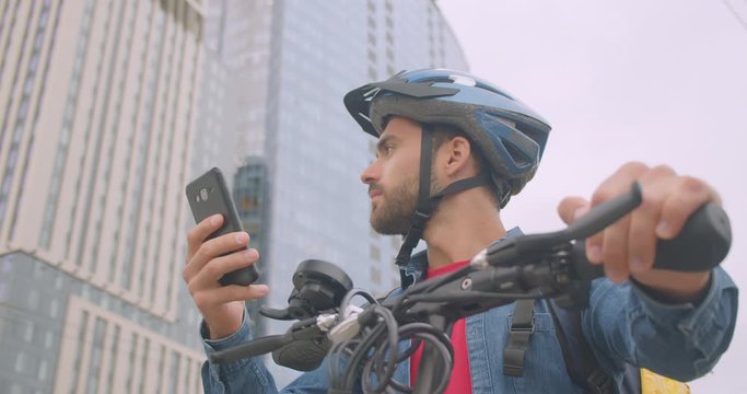 Closeup portrait of adult attractive caucasian delivery man standing with kick scooter and back pack using navigation app on phone in city
