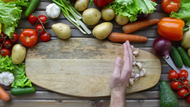 Top view of male hand moving garlic cloves to side of wooden cutting board with copy space; there is variety of fresh vegetables around on the table