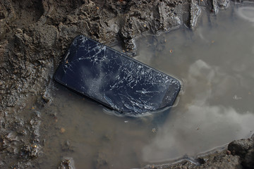 The smartphone with a broken display lies in a puddle and dirt, cracks and damage to the device.