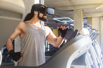 Closeup side view portrait of serious young adult sport man with long curly hair training alone. Handsome male doing exersices in a gym on treadmill and looking at vr glasses. Indoor, fitness concept