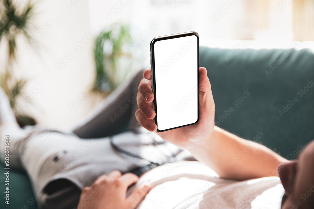 Poster Man using smartphone blank screen frameless modern design while lying on the sofa in home interior