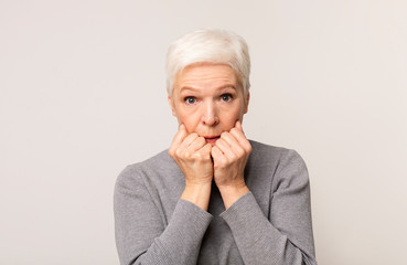 Scared elderly woman looking at camera, covering mouth with fists