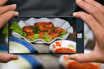 Woman photographing on cell phone grilled shrimps with salad on a plate and red sauce in a restaurant.