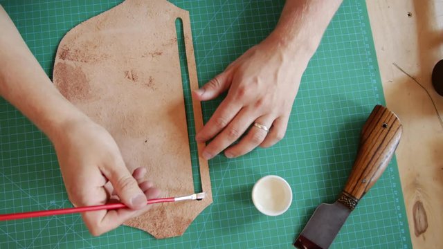 Top view of artisan hands holding brush and applying white glue on piece of leather