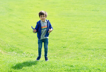 Fototapeta premium Full length portrait kid jumping in the green grass, Cute little boy playing plastic airplane toy, Child having fun playing outdoor in spring or summer field on sunny day.