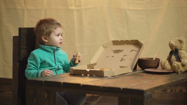  Boy eating pizza sitting at wooden table with toy bear. Funny boy and teddy bear eating pizza on burlap background. Tasty pizza. Burlap background. Junk food. Boy with bear and pizza. Unhealthy food.