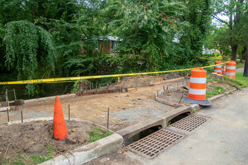Sidewalk in the process of being replaced. Damaged concrete blocks have been removed and wooden boards placed to construct forms to lay new concrete. There are orange barriers and caution tape at site