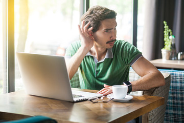 What? I can't hear you. Young businessman in green t-shirt sitting and try to listen something on laptop or video call. business and freelancing concept. indoor shot near big window at daytime.