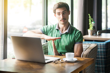 Time out! Young businessman in green t-shirt working, looking at laptop screen on video call and pleading to take more time. business and freelancing concept. indoor shot near window at daytime.