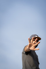 Bearded Man wearing a cap, view from the front, with the arm stretched to the camera with the palm of his hand opened covering is face partially. Blue sky.