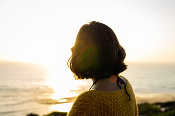 Girl with short hair and a yellow sweater watching the sunset on the beach below.