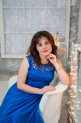 A very beautiful brown-haired woman with curly hair in a blue evening dress is sitting on a business sofa near the New Year's garland. Portrait, vertical format