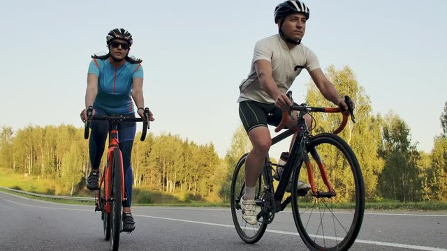 Steadicam shot of two healthy mem and woman peddling fast with cycling road bicycle at sunset.