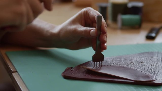 Close up of shoemaker hands holding line piercer and hammer making holes in piece of brown leather