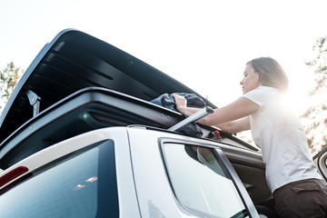 A woman puts things in the roof rack of a car or in a cargo box, before a family trip on vacation, against the sky and trees, on a summer evening.