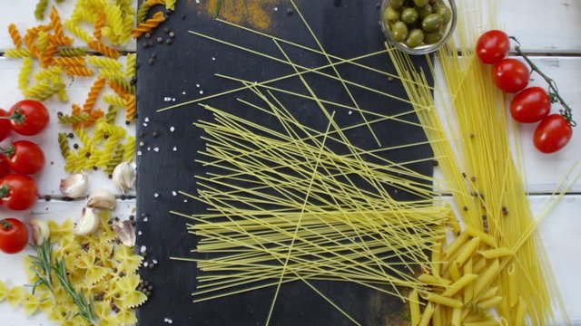 Top view of hand of cook throwing bunch of spaghetti on kitchen table with different types of Italian pasta, olives, plum tomatoes, garlic cloves, peppercorns and seasonings on it