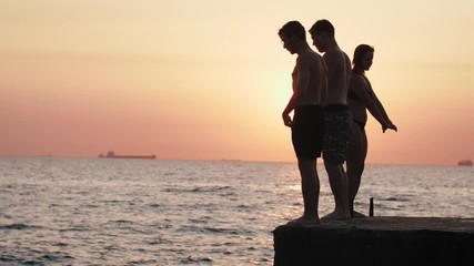Group of young friends jumping and doing tricks from a pier into the sea during beautiful sunrise, slow motion