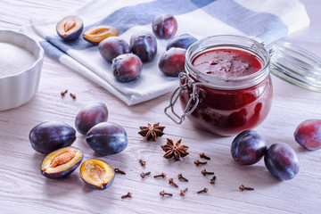 An open glass jar with homemade plum jam, fresh ripe plums, folded towel, ceramic bowl with sugar, badyan seeds and dry carnation grains on a wooden table in the morning light