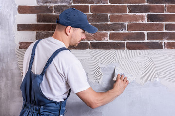 A worker in blue overall and cap, holding a comb putty-knife and covering a wall surface with...