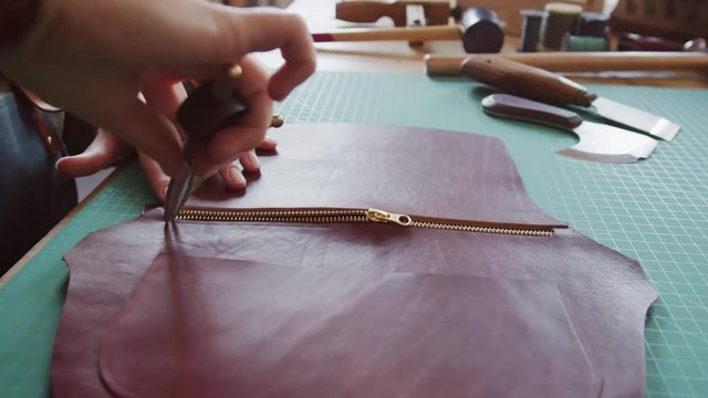 Close up of female artisan holding special steel tool, gluing zipper and piece of brown raw leather and then checking zipper