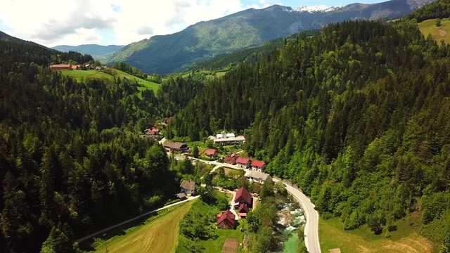 Forward drone shot, above Luče, Slovenia, next to a flowing river, surrounded by mountains covered in bright green forests, on a sunny day