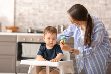 Mother giving her little son nibbler with tasty food in kitchen at home