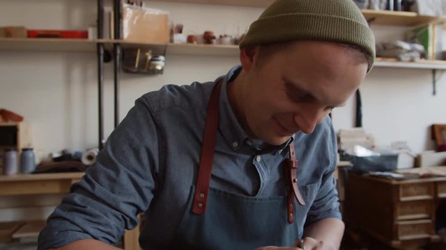 Tilt down of tanner sitting at workbench and making holes in leather product using small line piercer and hammer