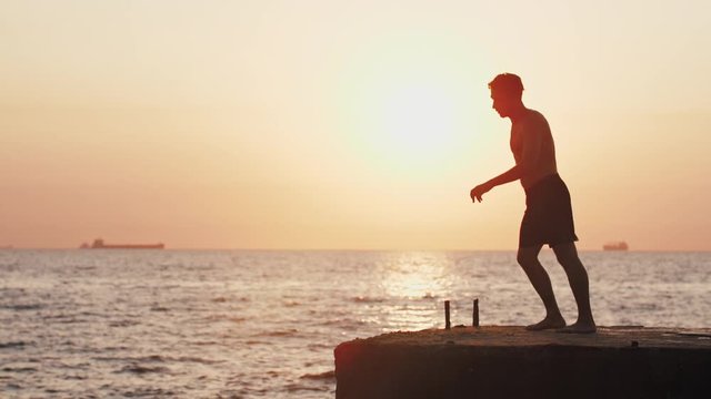 Young man jumping from sea pier and doing backflip during beautiful sunrise, super slow motion