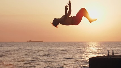 Young man jumping from sea pier and doing backflip during beautiful sunrise, super slow motion