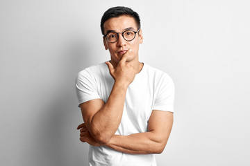 Smart Kazakh dressed in casuals and glasses thinks and doubts, holds his chin with hand on white studio background. Responsible Asian makes a decision