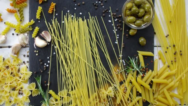 Top view of bunch of spaghetti being thrown on kitchen table with different types of Italian pasta, olives, garlic cloves, peppercorns and seasonings on it
