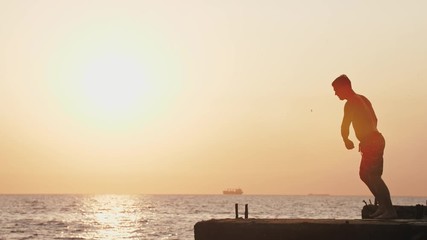 Young man jumping from sea pier and doing backflip during beautiful sunrise, super slow motion