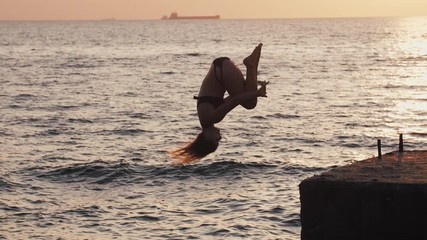 Young youman jumping from sea pier and doing frontflip during beautiful sunrise, slow motion