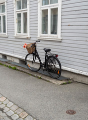 Fototapeta na wymiar A black fashionable bicycle with a basket on the handlebars stands against the wall of an old wooden white house on a paved sidewalk