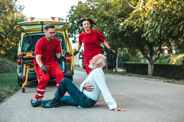 Elegant senior man with heart attack symptoms sitting on the road emergency medical service workers trying to help him. Driver assistance service.