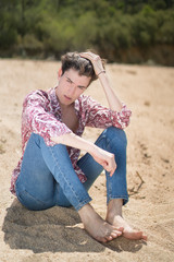 young man sitting on the sand with jeans and flower shirt with fixed look