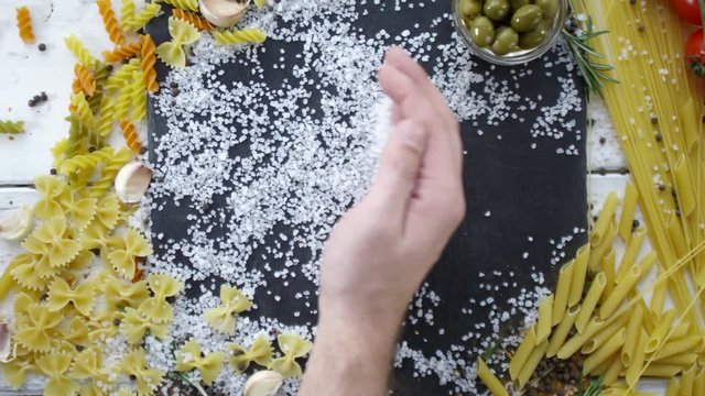 Top view of male hand moving salt crystals to the side of black cutting board with variety of Italian pasta, olives, garlic cloves, tomatoes and seasonings around. Copy space in center