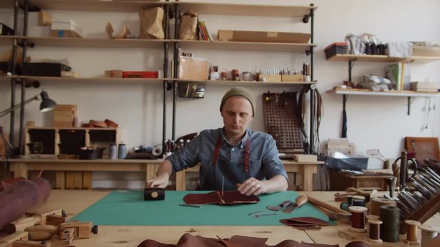Zoom in of artisan sitting at worktop in tannery and making holes in leather using line piercer and hammer