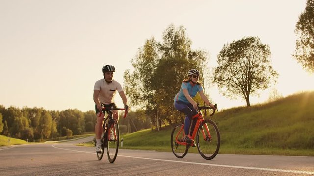 Steadicam shot of two healthy mem and woman peddling fast with cycling road bicycle at sunset.