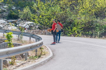 Two male longboarders carrying their longboards in their hands while climbing uphill and preparing for a downhill slide. Wearing red t-shirts, green hat, and super cool sunglasses.