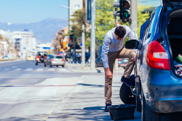 A strong businessman is bending his knee while trying to change a flattie on his car with his lug wrench.