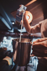 Talanted barista is pouring milk to the jug for latte or cappuchino at his cafe.